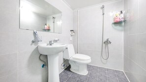 Een badkamer met een witte wastafel, toilet en douche. Boven de wastafel hangt een spiegel en er is een plank met toiletartikelen.