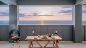 A dining table with food and wine glasses, a grill, and a view of the ocean at sunset.