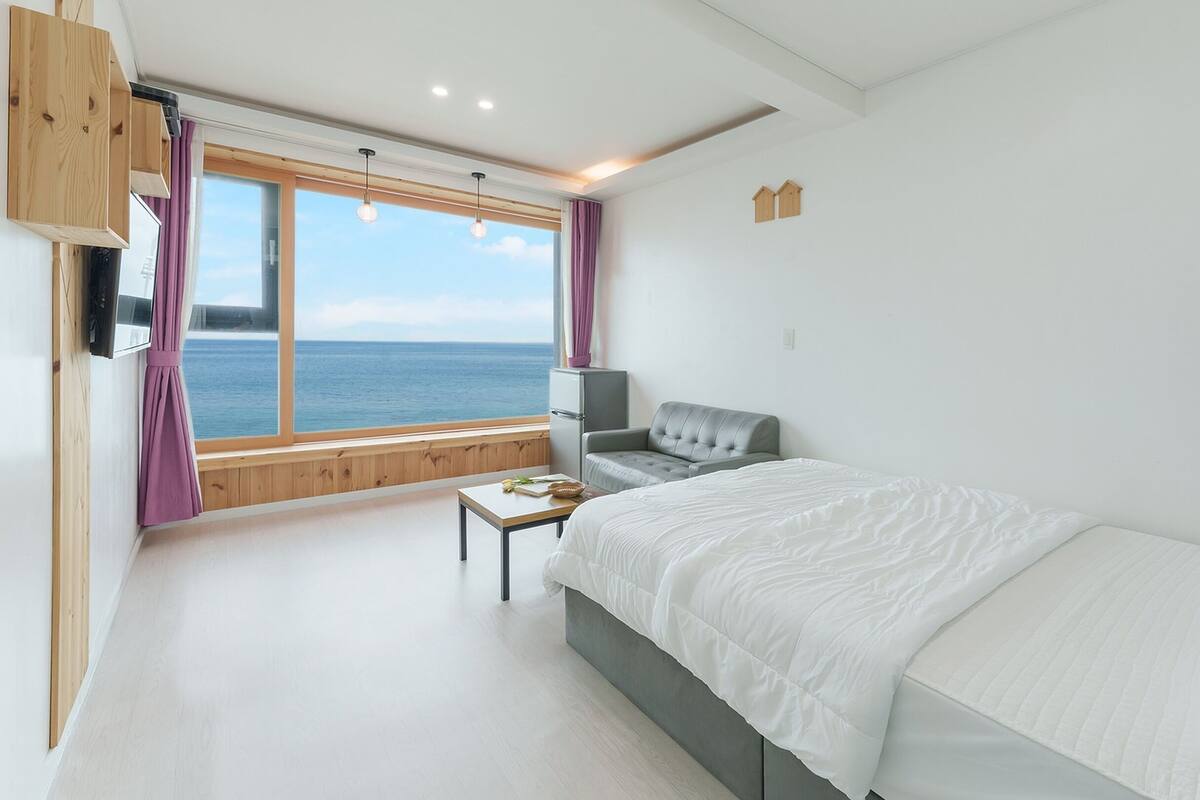 Een slaapkamer met een groot raam met uitzicht op zee, een bed met wit beddengoed, een grijze bank en een klein tafeltje met een boek en bloemen.