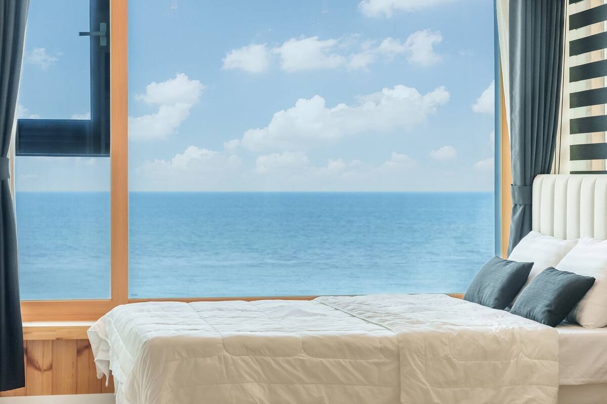 Een slaapkamer met een bed, uitzicht op zee en een raam met gordijnen.