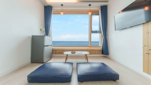 Quarto compacto com uma grande janela, TV de tela plana, pequena mesa e duas áreas de assento acolchoadas em azul.