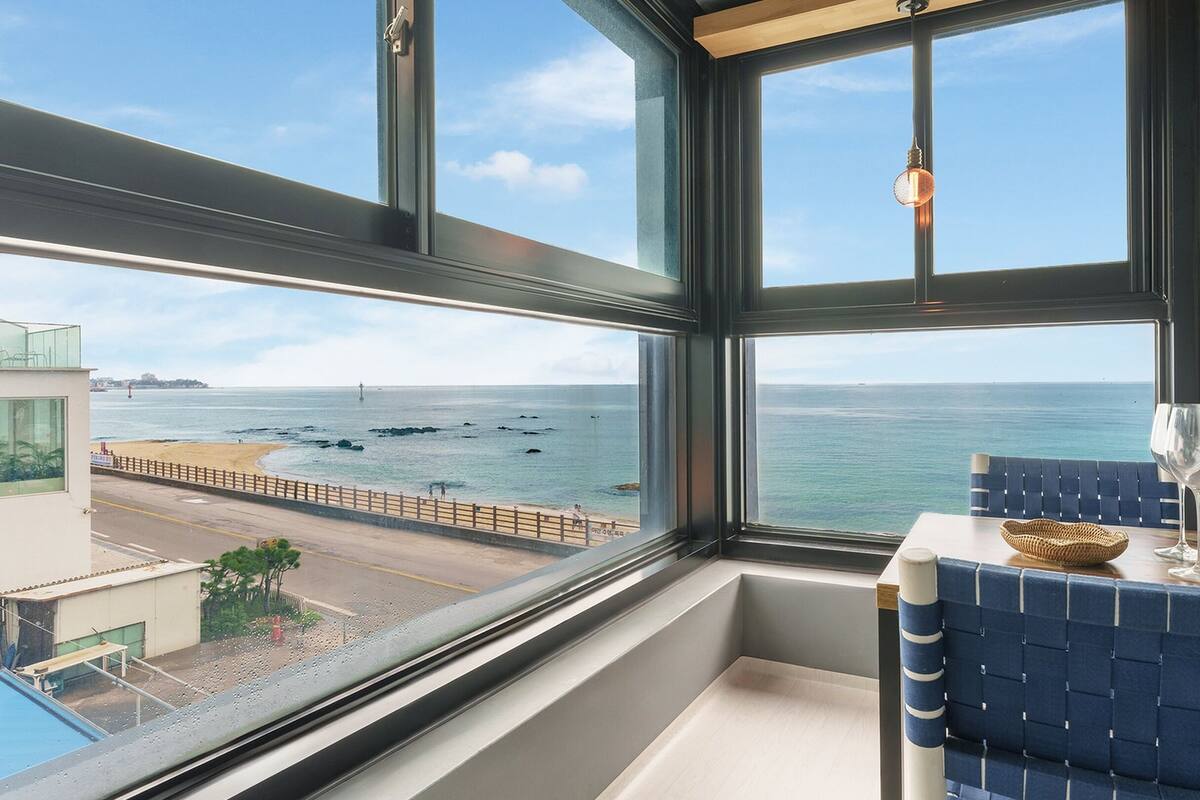 Een kamer met een groot raam met uitzicht op zee, een klein tafeltje met een mandje, en een blauwe stoel.