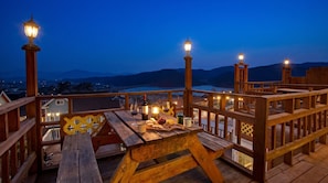 Una terraza de madera con una mesa dispuesta para comer, con vistas a un paisaje montañoso y una masa de agua.