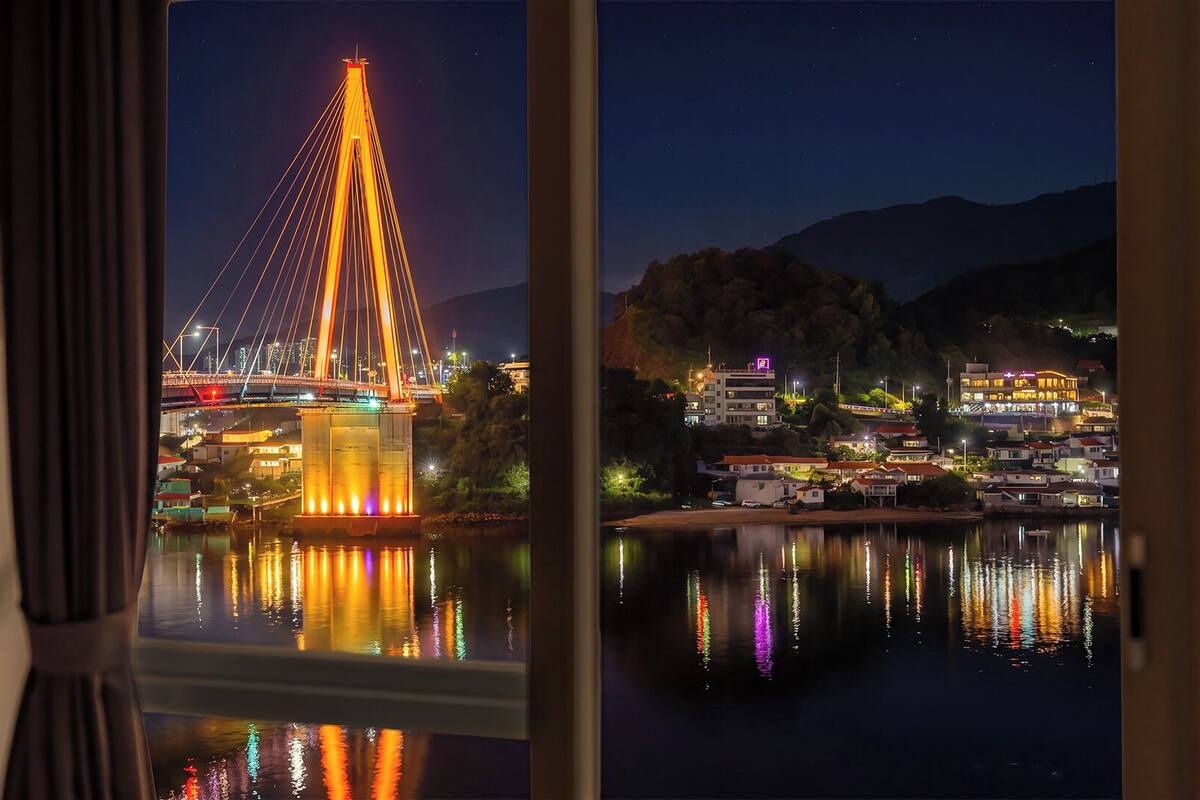 Une vue nocturne d’un pont illuminé, d’une étendue d’eau reflétant les lumières et d’une ville avec des bâtiments en arrière-plan.