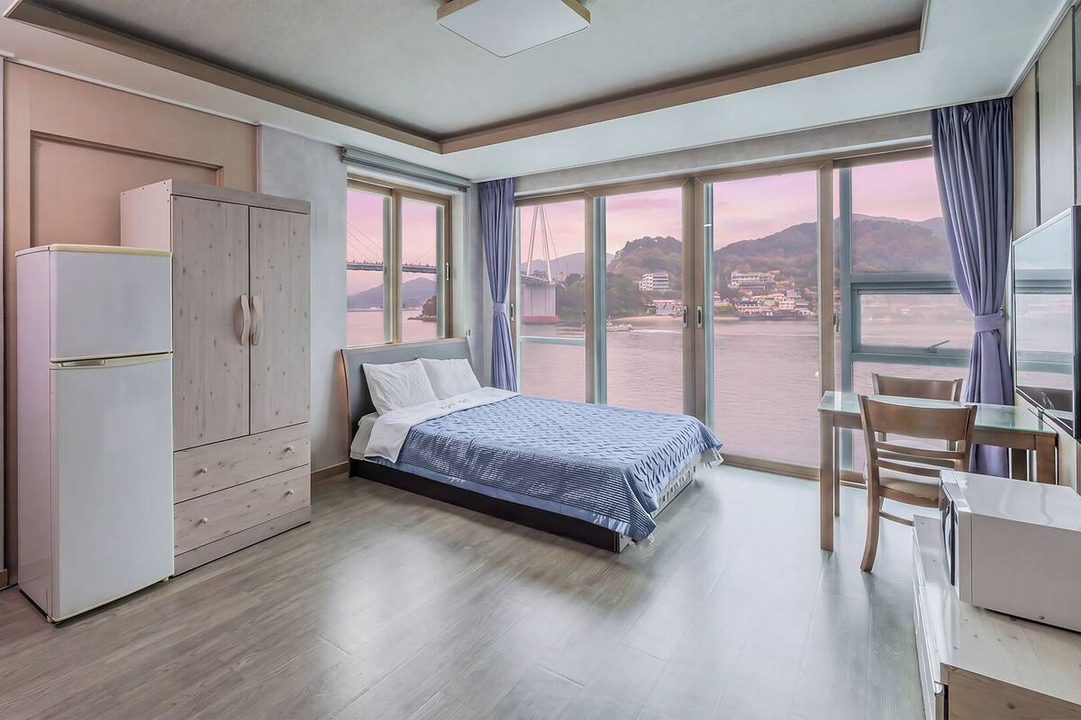 Une chambre à coucher comprenant un lit, un bureau, une chaise, une armoire et de grandes fenêtres donnant sur un pont et des collines.