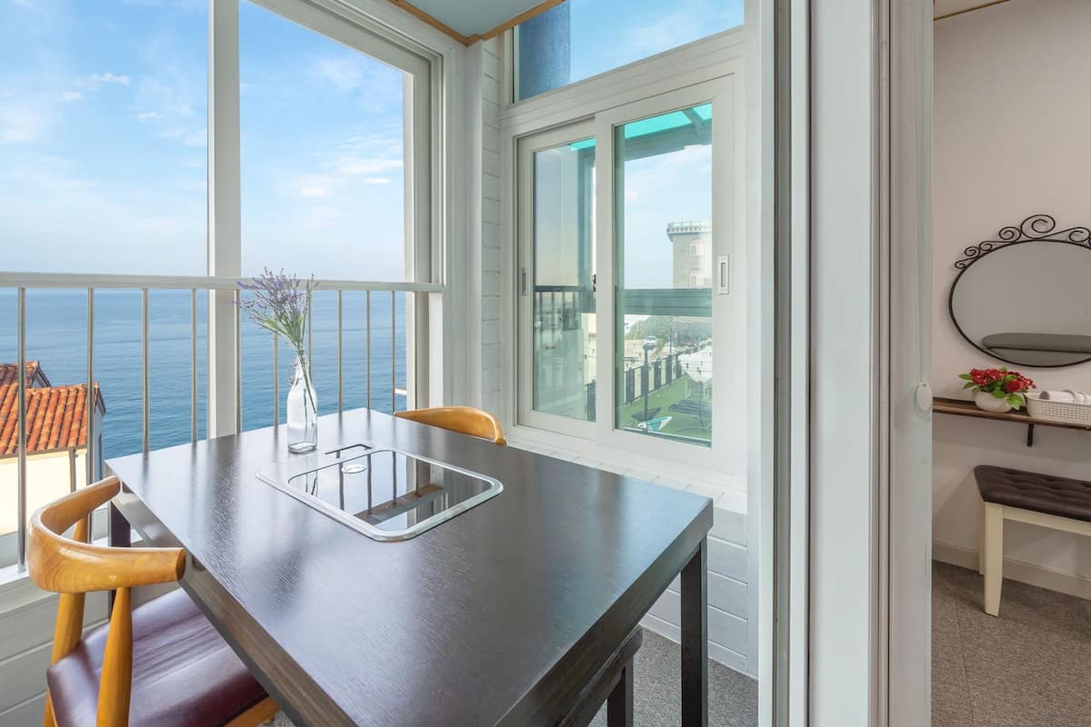A balcony with a table and chairs, a vase with flowers, and a view of the sea and buildings.