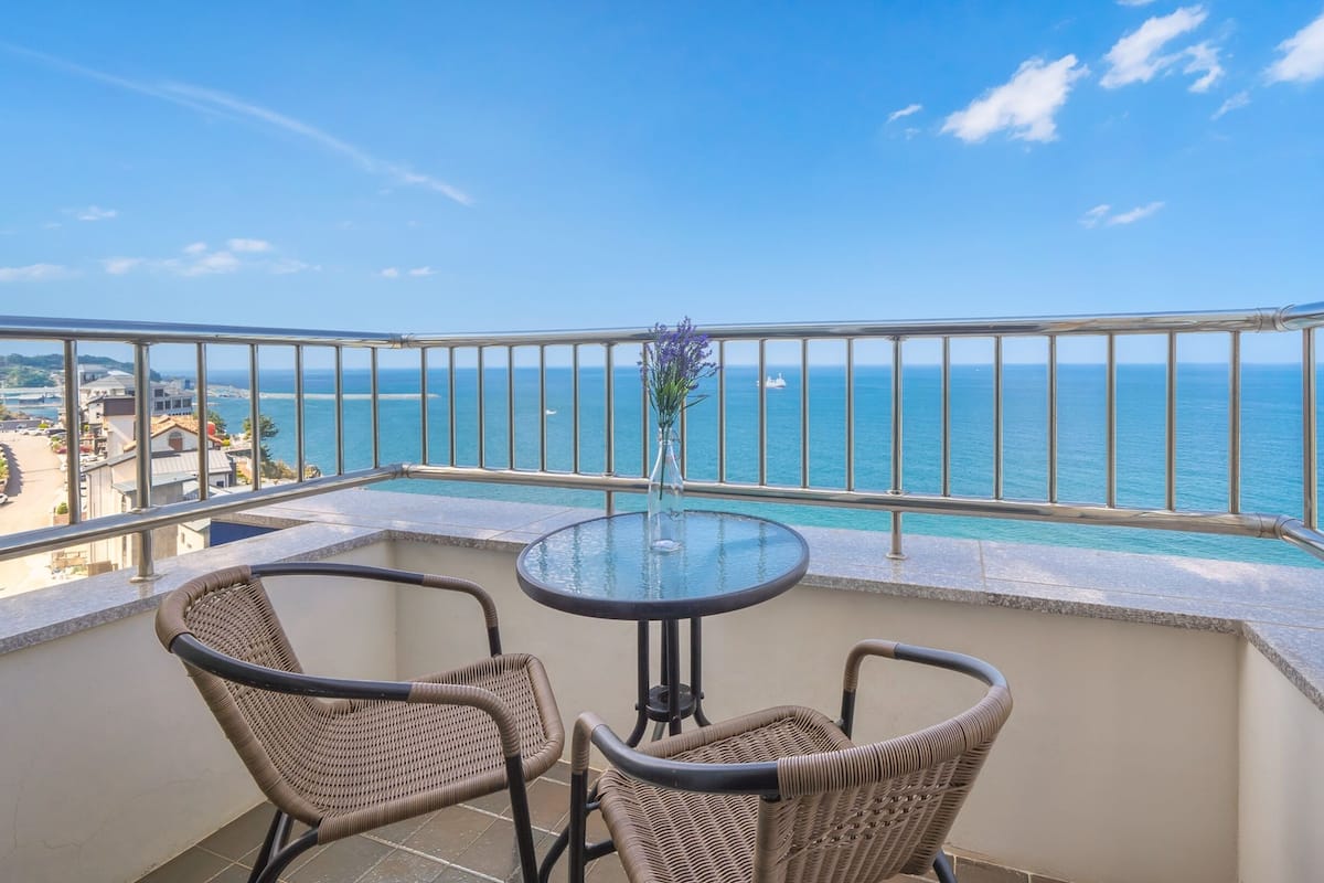 A balcony with wicker chairs and a glass table overlooking a coastal view.