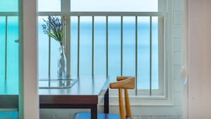 Una habitación con vista al mar a través de una ventana con persianas blancas. Hay una silla de madera y una mesa con un florero.