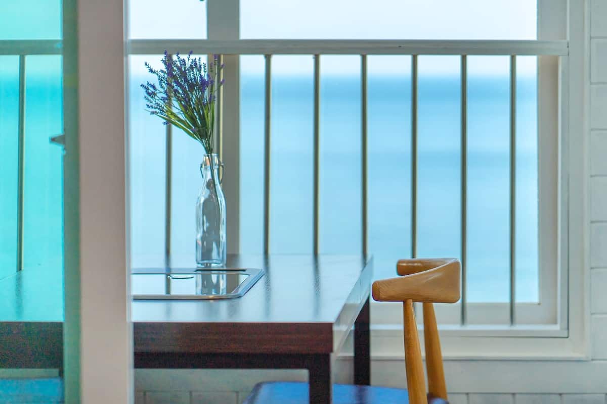 A room with a view of the sea through a window with white blinds. There is a wooden chair and a table with a vase of flowers.