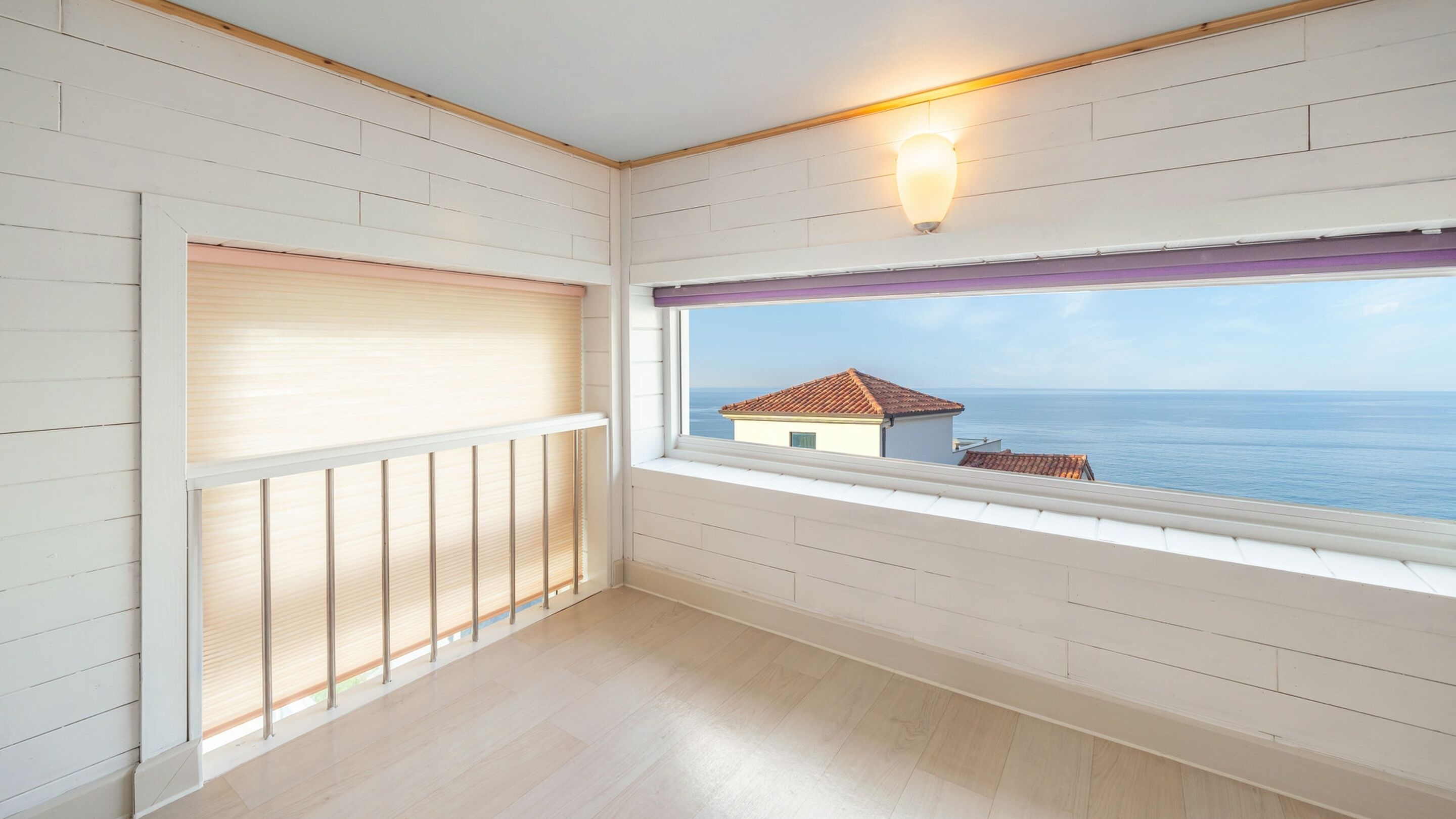 A balcony with a view of the sea, a window with blinds, and a wall-mounted light fixture.