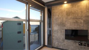 A modern interior with a large sliding glass door leading to a balcony with a view of the sea and buildings. A flat-screen TV is mounted on the wall. - Donghae Haru Pension (Donghae)