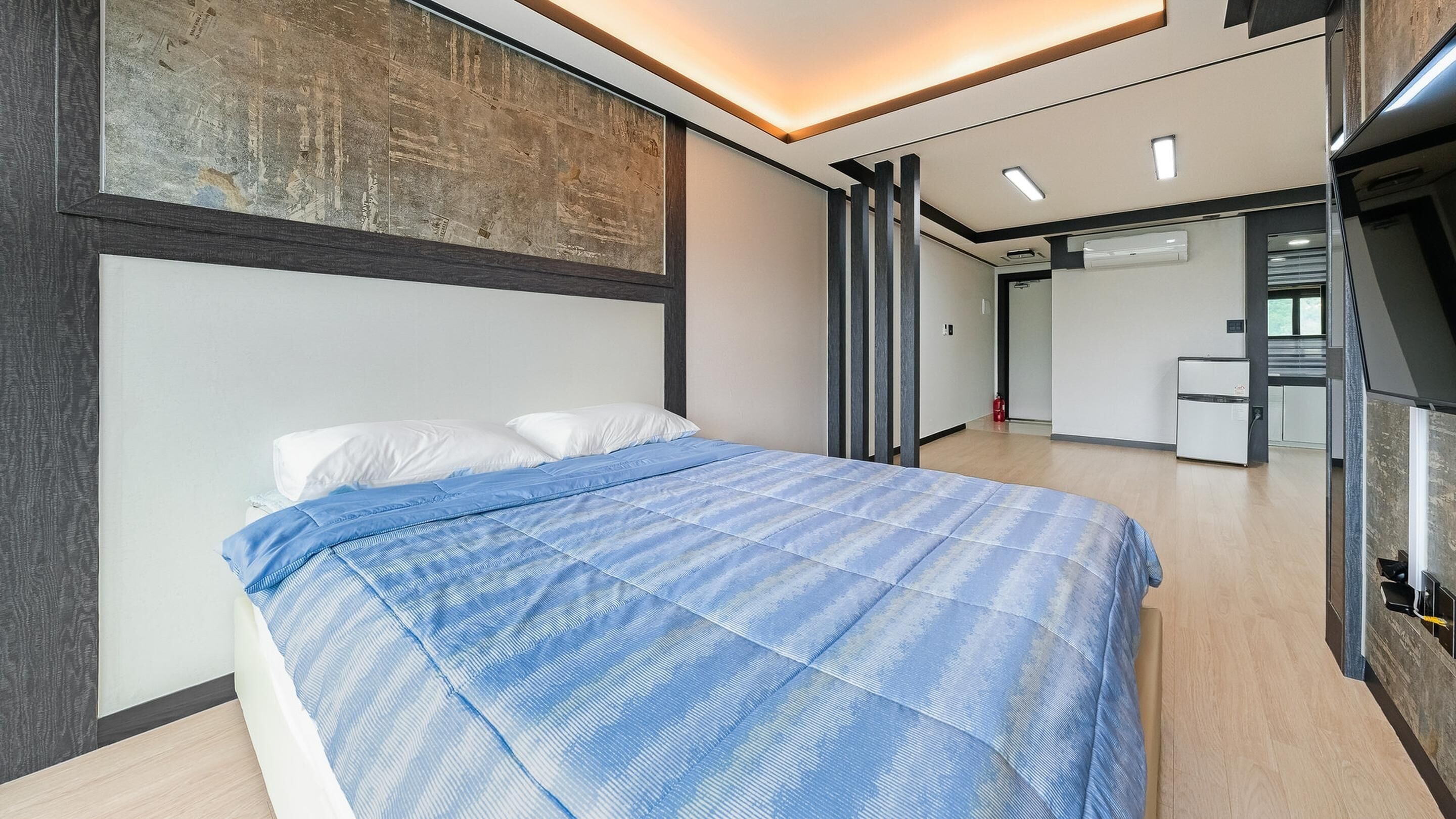 A modern bedroom with a bed, a large artwork on the wall, and a kitchenette in the background.
