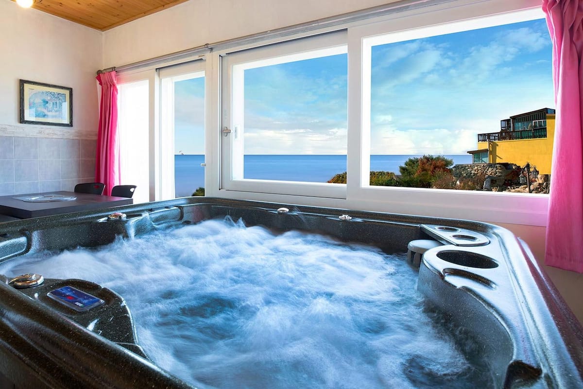 Un bain à remous avec vue sur l’océan, une table à manger, des chaises et des rideaux roses.