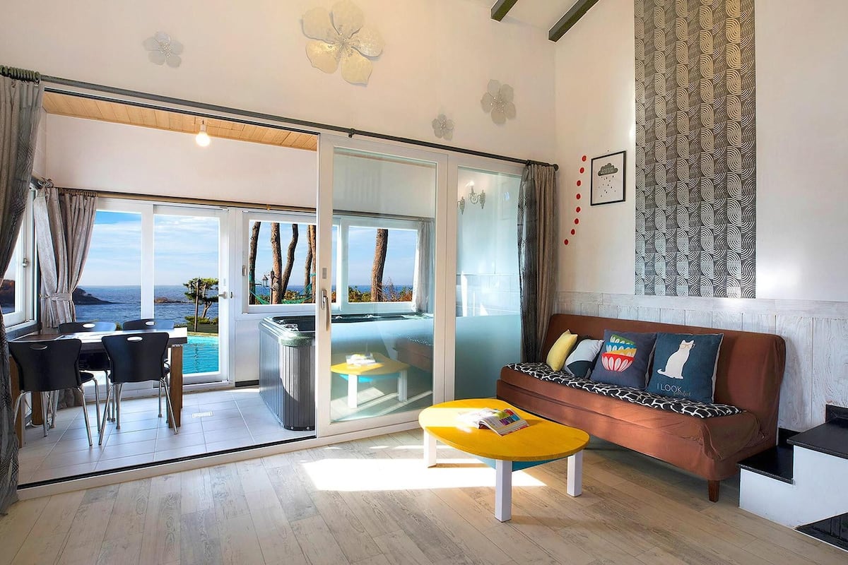 Un salon moderne comprenant un canapé, une table basse et un coin repas avec des chaises. Une porte-fenêtre coulissante donne accès à un balcon offrant une vue sur la mer.