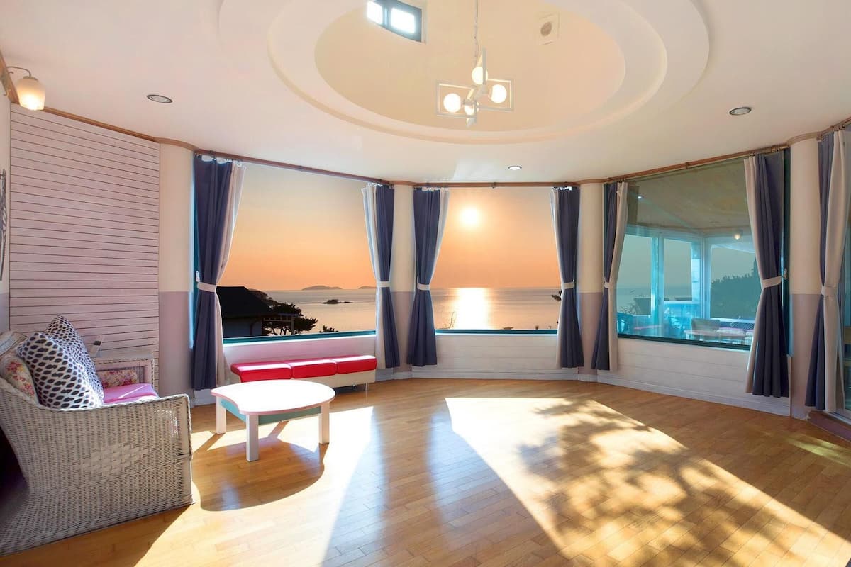 Un salon moderne doté d’une grande fenêtre donnant sur la mer, d’un parquet, d’une table ronde et d’un canapé en osier.