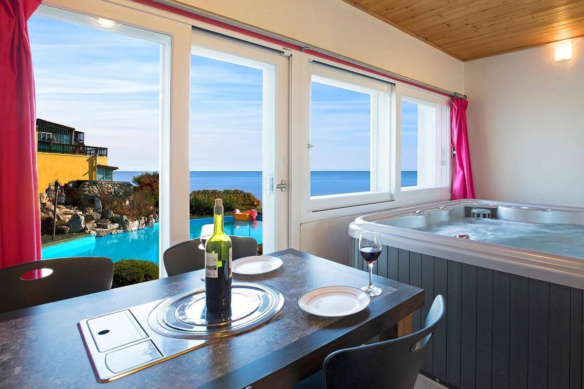 Un balcon avec un jacuzzi, une table avec une bouteille de vin et une vue sur une piscine et l’océan.