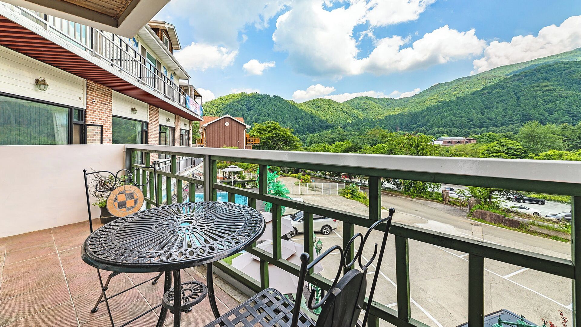 A balcony with a metal table and chairs, overlooking a parking area and lush green hills.