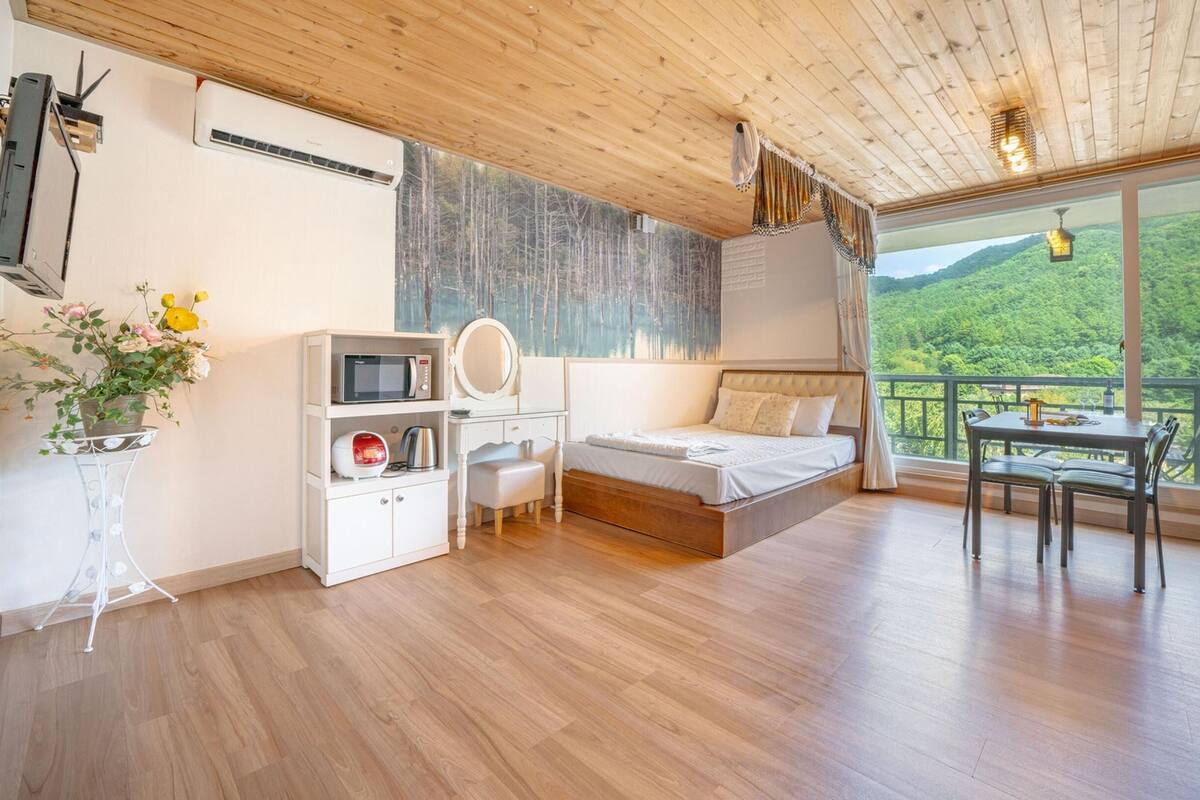 Ein Schlafzimmer mit Bett, Nachttisch und Blick auf einen Wald durch ein großes Fenster.