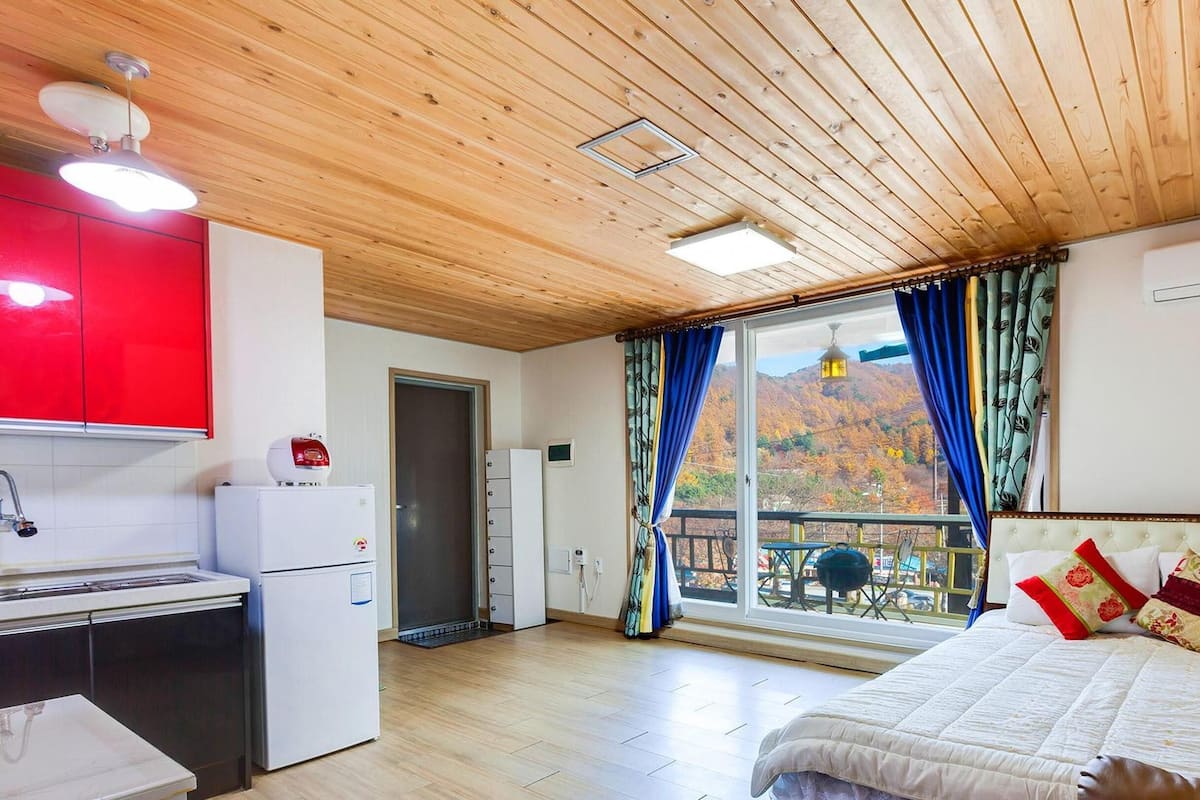 Ein Schlafzimmer mit Bett, Schreibtisch, einem roten Schrank, einem weißen Kühlschrank und einem Fenster mit Vorhängen.