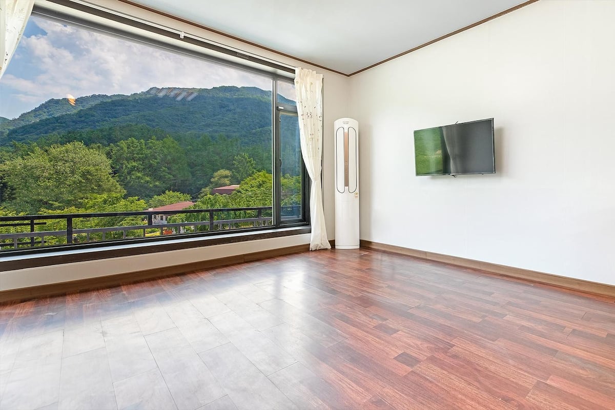 Ein Zimmer mit großem Fenster, Holzboden, Heizlüfter und Wandfernseher.