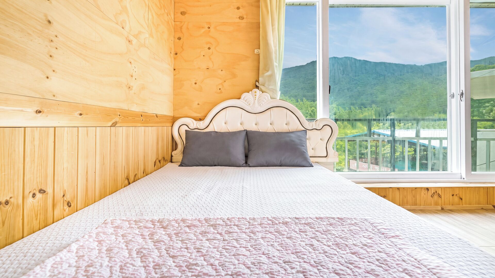A wooden room with a bed, a window, and a view of green hills.