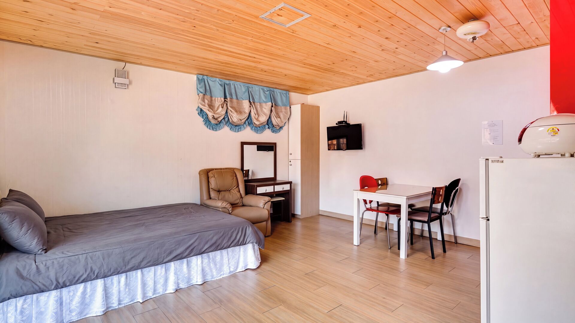 A bedroom with a bed, a desk, and a chair. There is a mirror on the wall, a ceiling light, and a refrigerator.
