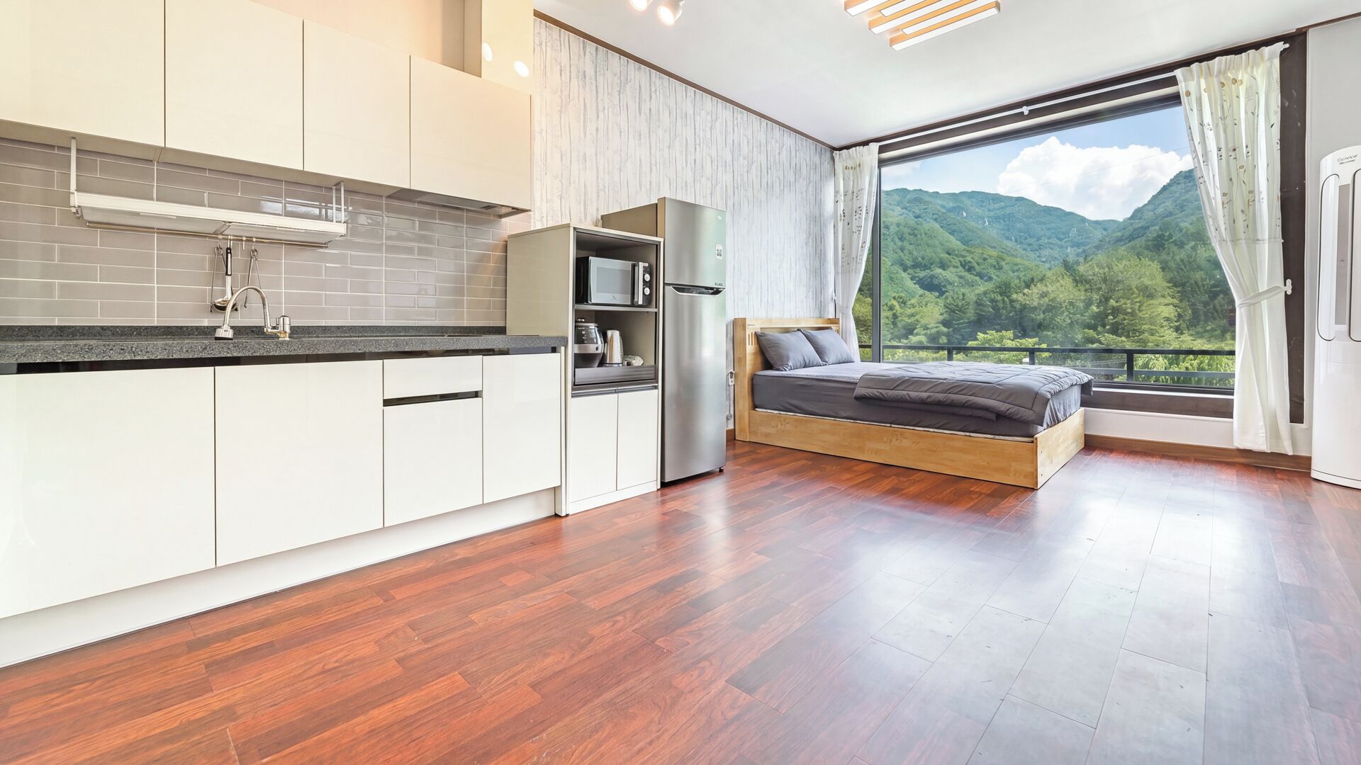 A modern bedroom with a wooden bed, a kitchen with white cabinets, and a large window with a mountain view.