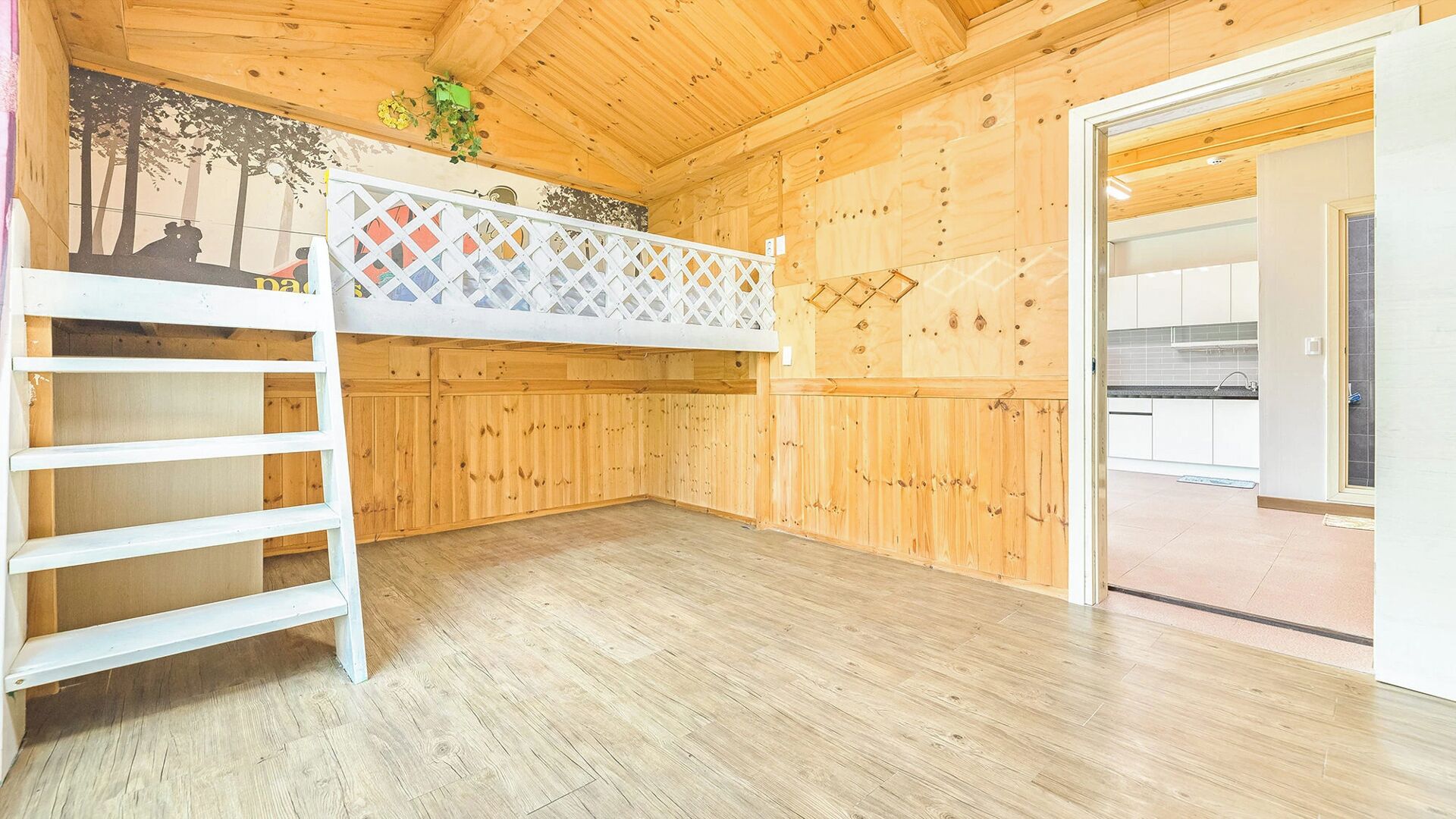 A wooden interior with a bunk bed, a ladder, and a kitchen area visible through an open door.