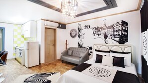 A bedroom with a bed, a grey armchair, a wall with a cityscape mural, a chandelier, and a small kitchenette. - Geoje Badasunjang Pension (Geoje)