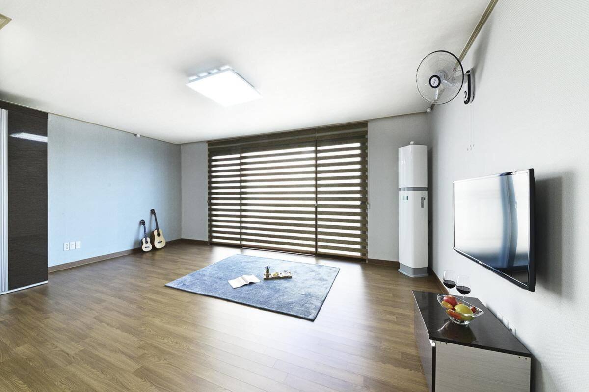 Un salon moderne avec un plancher en bois, une grande fenêtre équipée de stores, un téléviseur à écran plat fixé au mur et une petite table sur laquelle se trouve un saladier rempli de fruits et quelques verres.