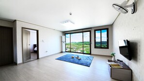 Ein geräumiges Zimmer mit großem Fenster und Blick auf eine grüne Landschaft, einem blauen Teppich und einem Flachbildfernseher auf einem Holzständer.
