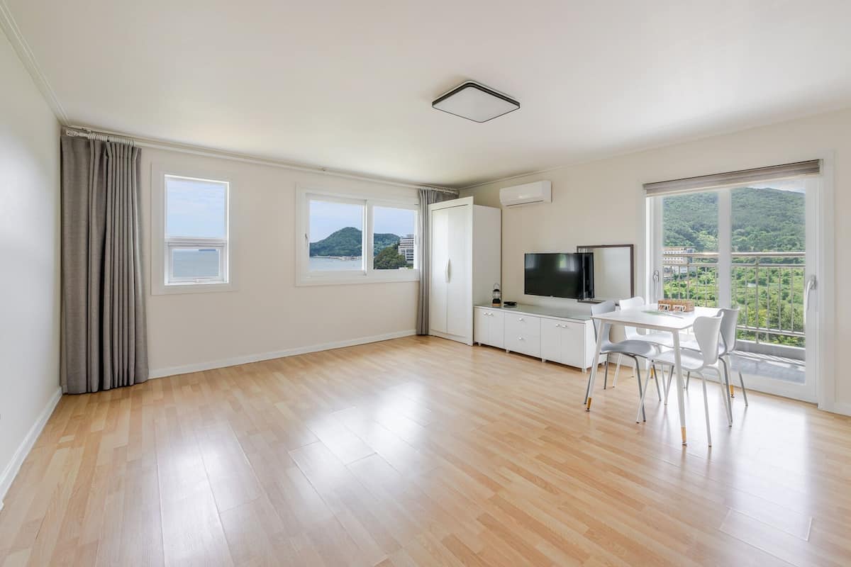 A spacious living area with a wooden floor, a dining table with chairs, a flat-screen TV, and large windows offering a view of the sea and greenery.