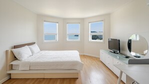 Een slaapkamer met een bed, een televisie, een spiegel en drie ramen met uitzicht op zee.