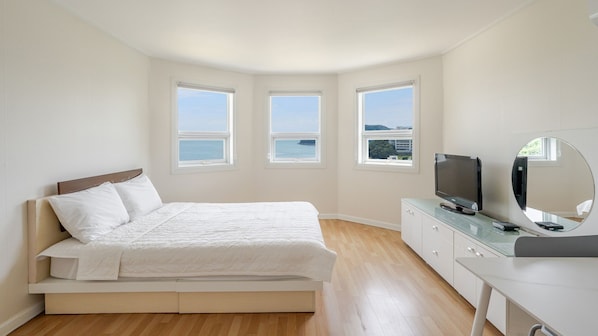 Un dormitorio con cama, televisión, espejo y tres ventanas con vistas al mar.