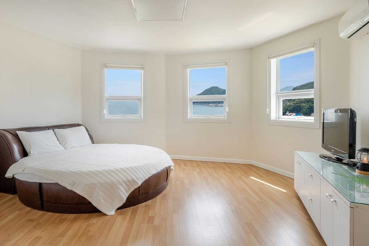 A bedroom with a bed, two windows with a view of the sea, a TV on a cabinet, and an air conditioning unit.