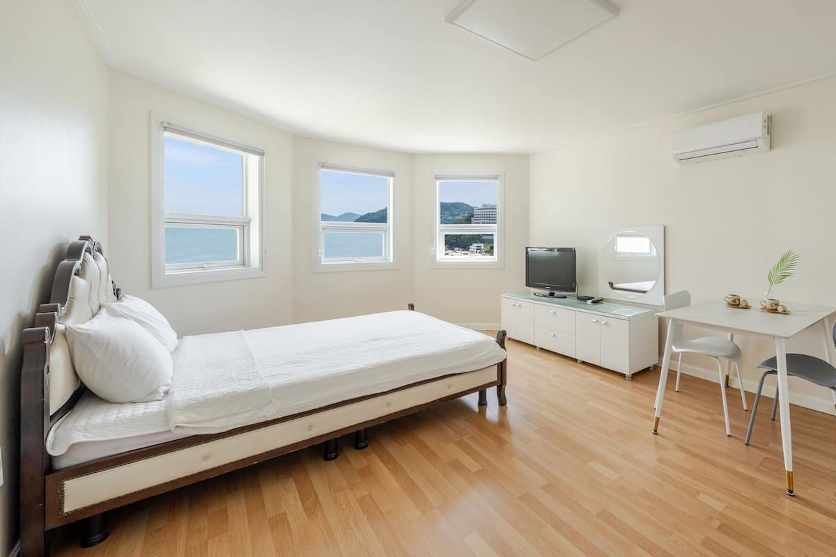 A bedroom with a bed, a desk, a chair, a television, and a window with a view of the sea.