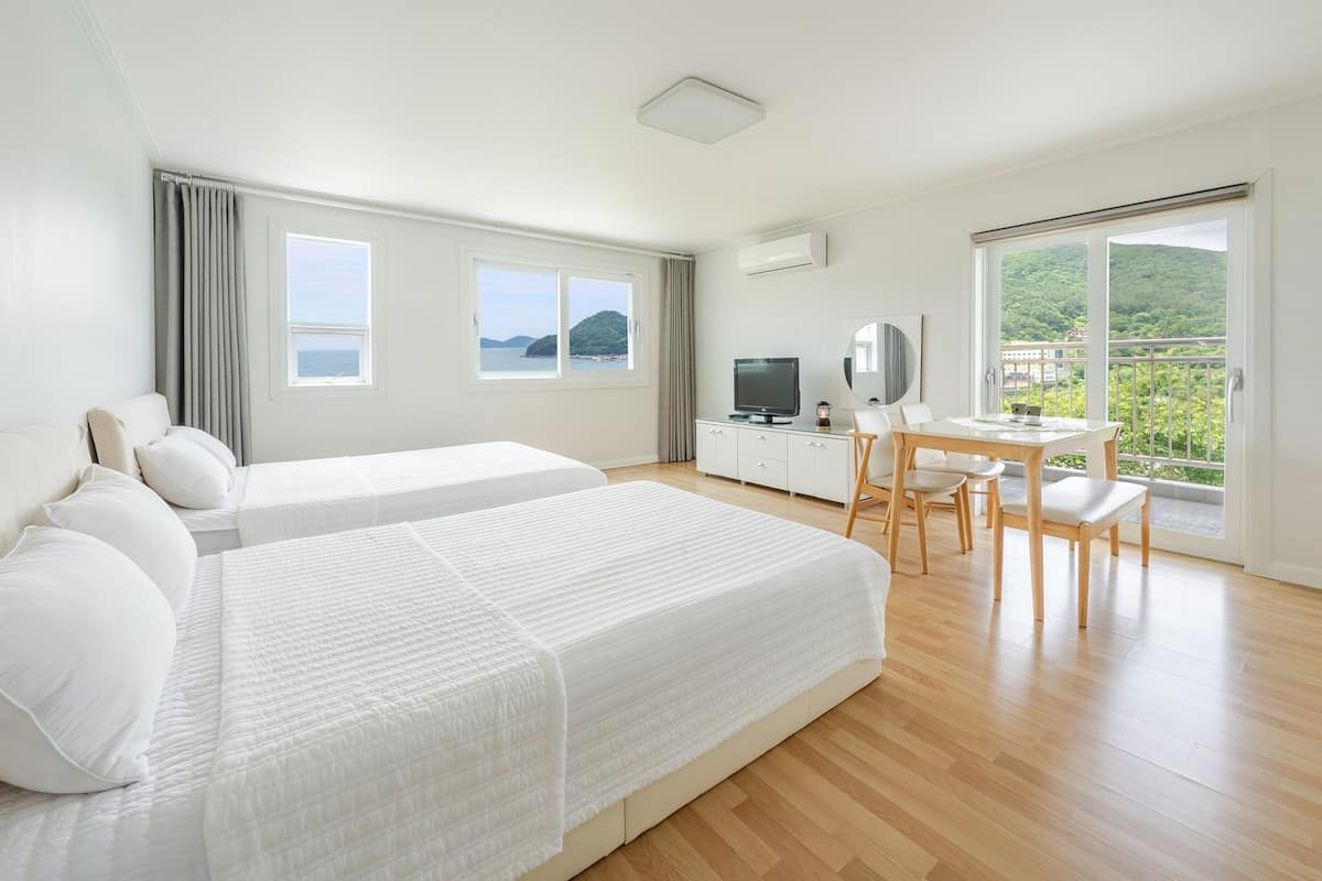 A hotel room with two beds, a desk with a chair, a TV, and a balcony with a view of greenery.