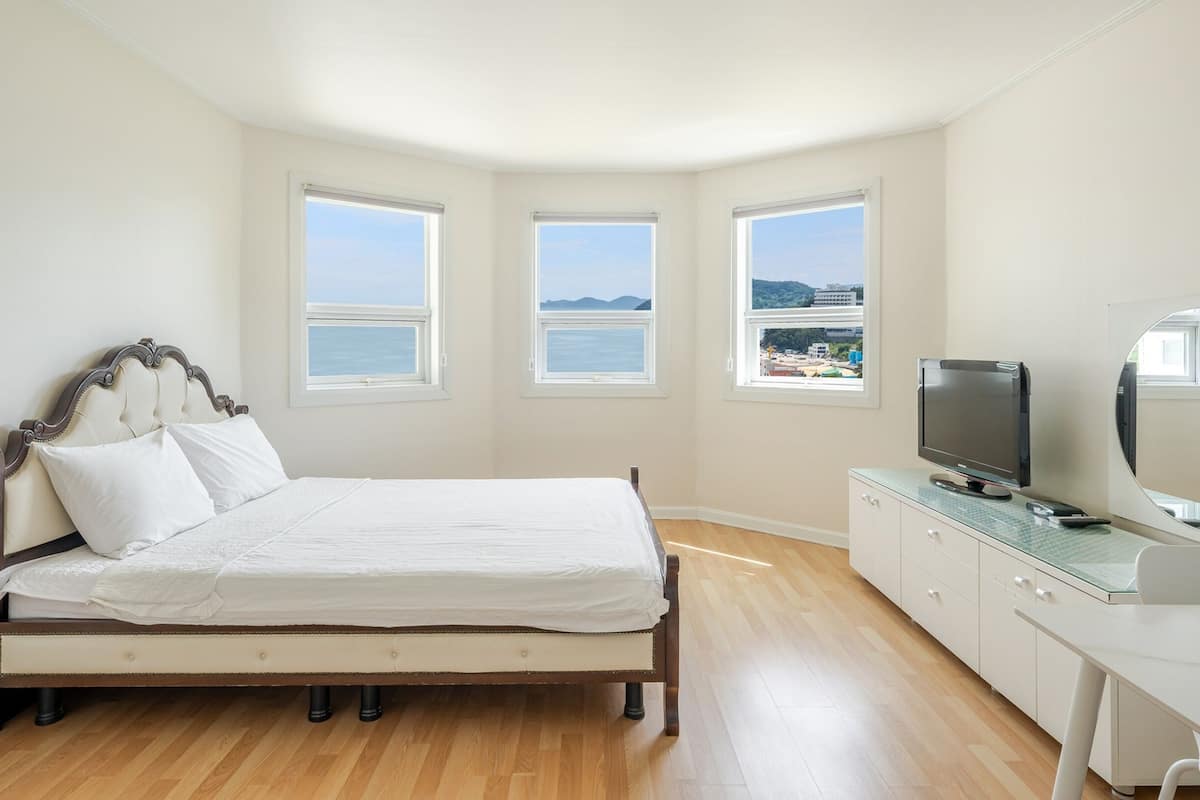 A bedroom with a bed, a TV, a desk, and three windows with a view of the sea.