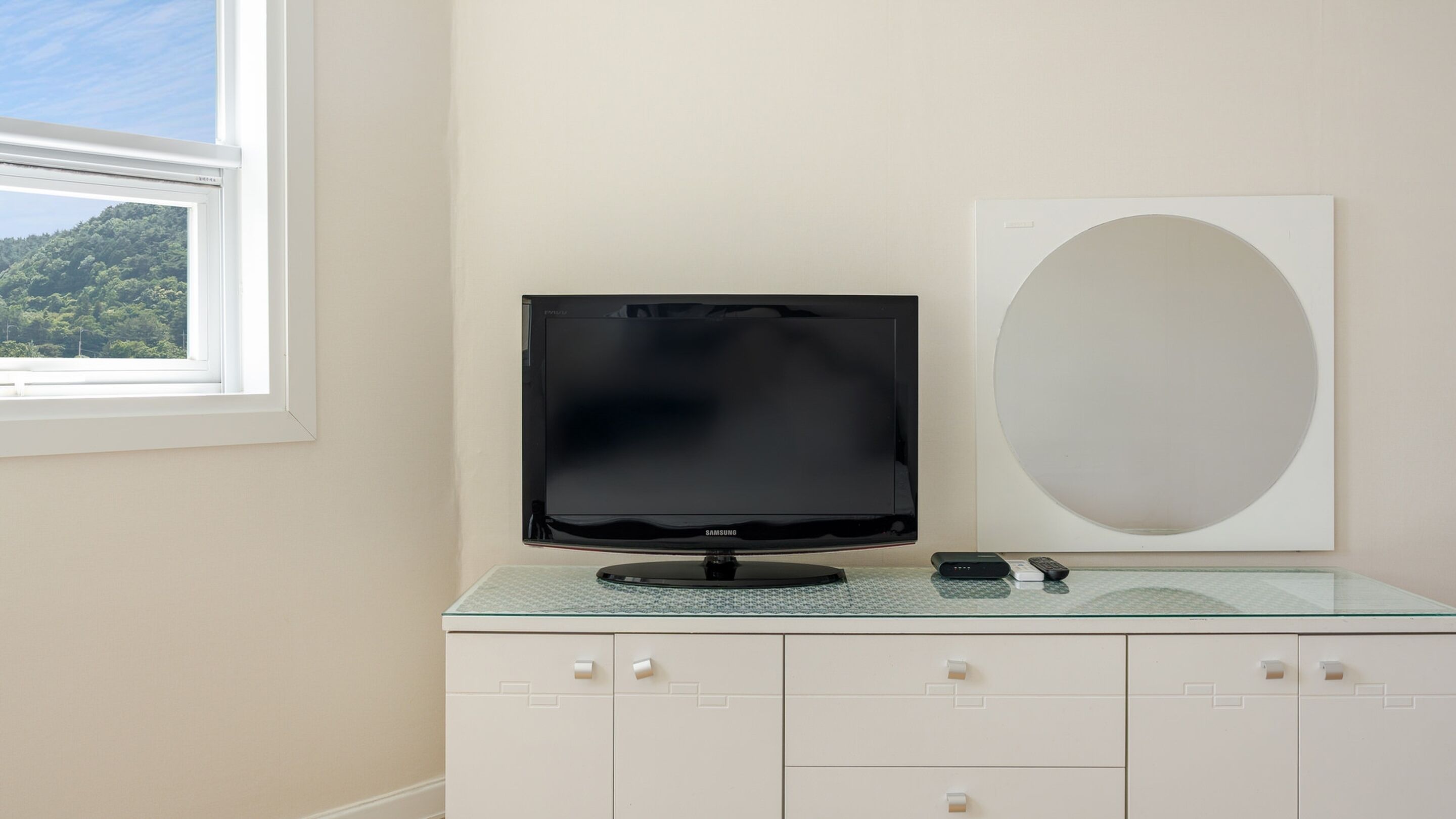 Una habitación moderna con una televisión de pantalla plana sobre un mueble de vidrio, un espejo ovalado grande y una ventana con vistas a un bosque.