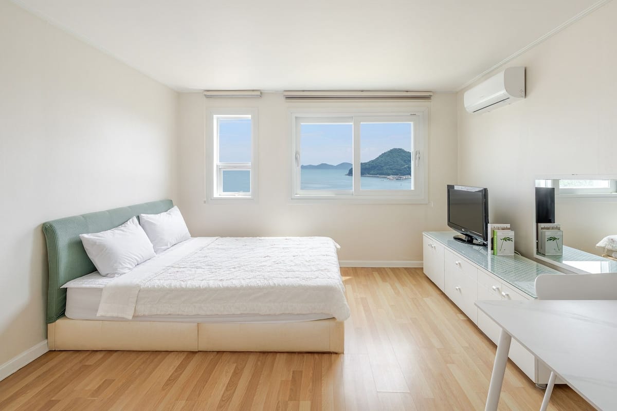 A bedroom with a bed, a TV, a desk, and a window with a view of the sea.
