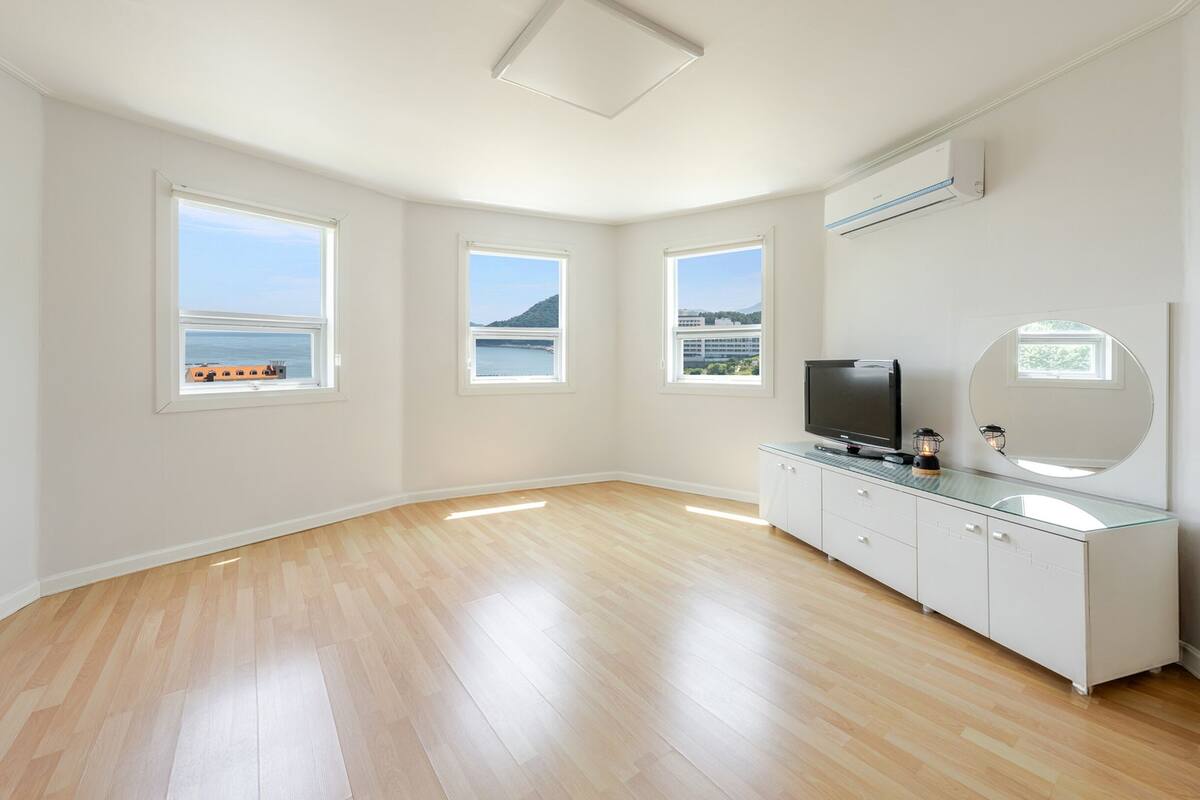 A spacious room with a wooden floor, white walls, and large windows offering a view of the sea.