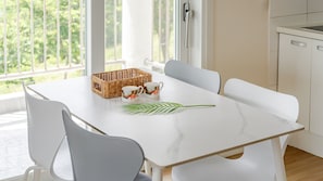 Una mesa de comedor con sillas blancas, una cesta de mimbre, dos tazas con motivos florales y una hoja verde.