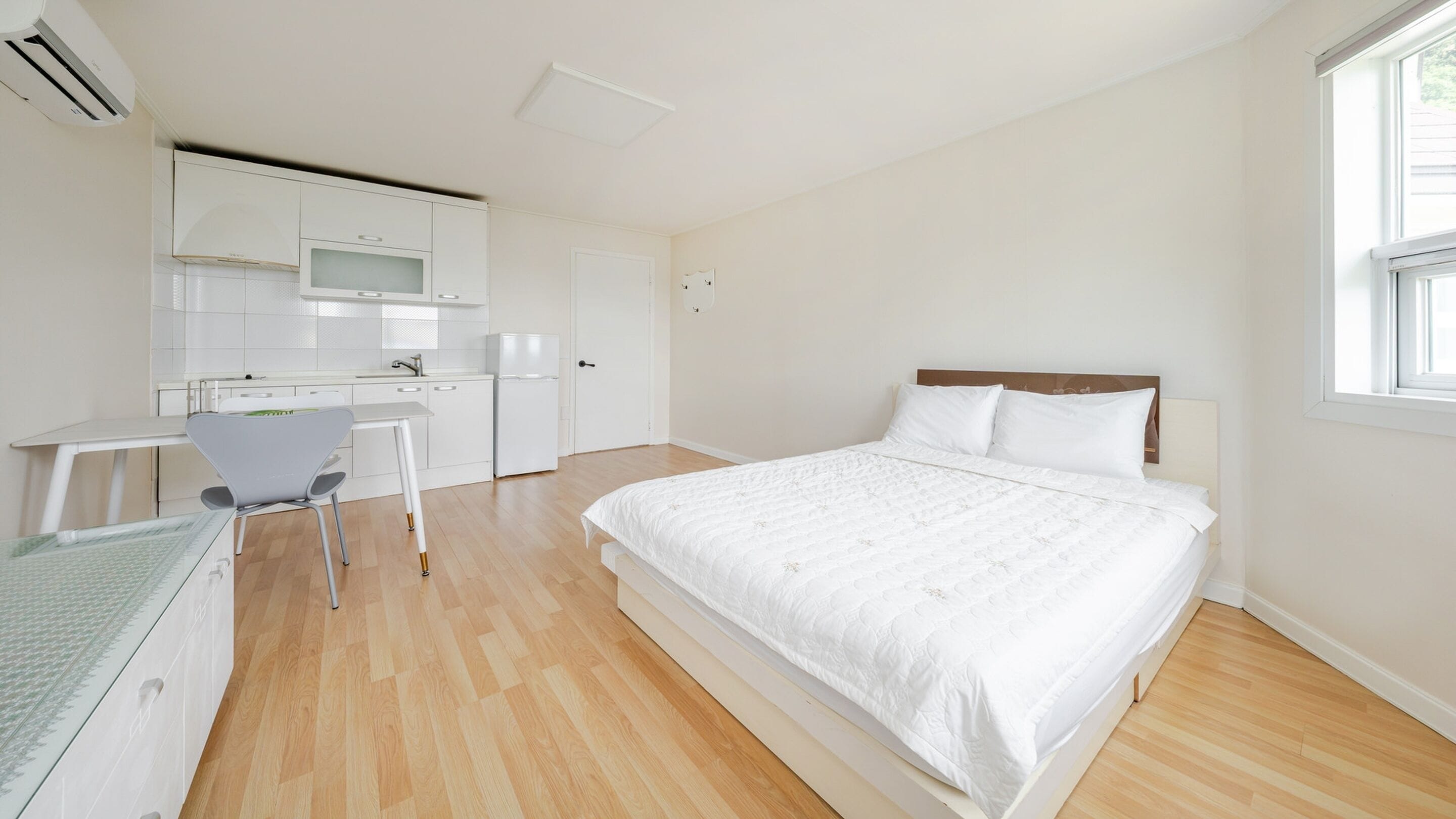 A bedroom with a large bed, a desk with a chair, and a kitchenette in the background.