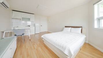 A bedroom with a large bed, a desk with a chair, and a kitchenette in the background.