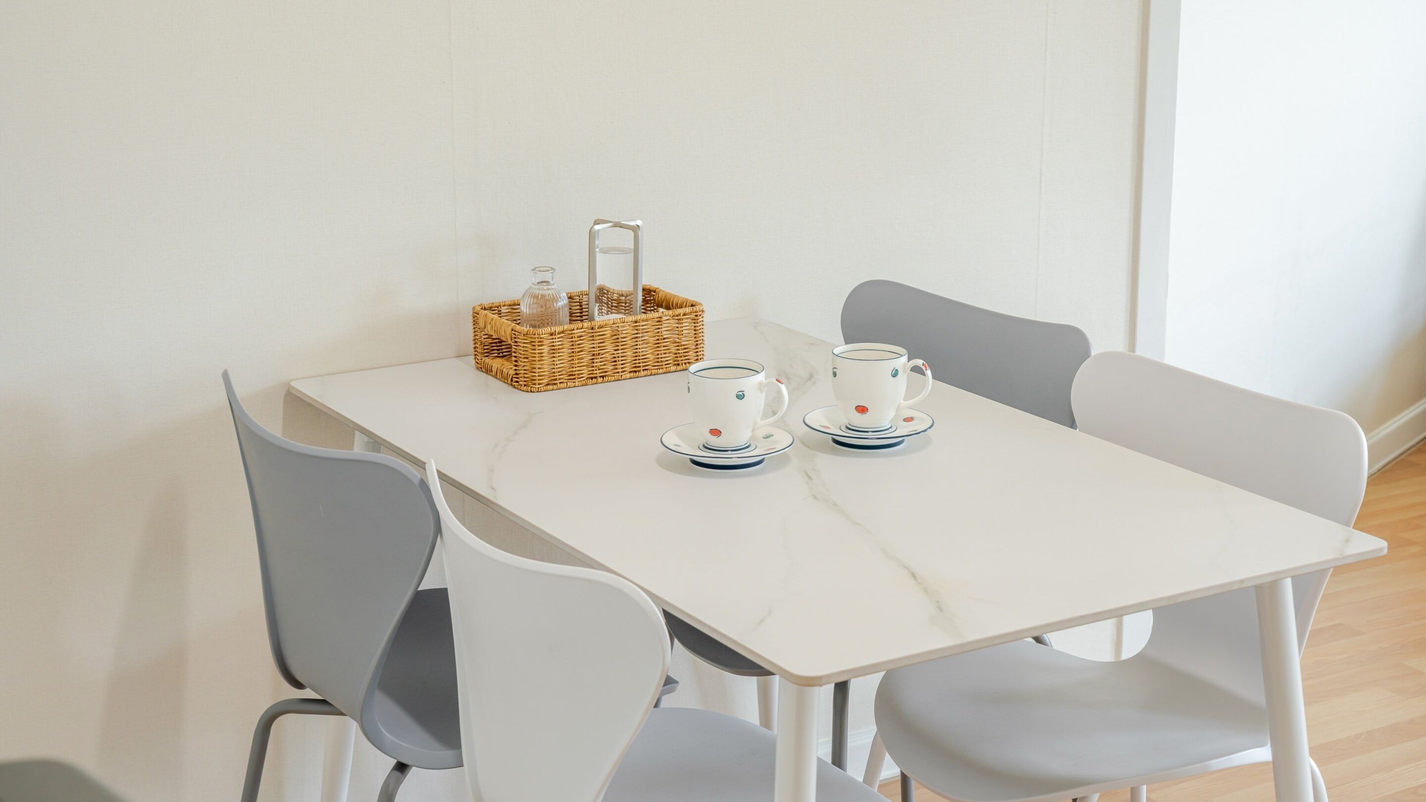 Une table blanche avec deux chaises blanches, un panier en osier et deux tasses à thé blanches avec leurs soucoupes.