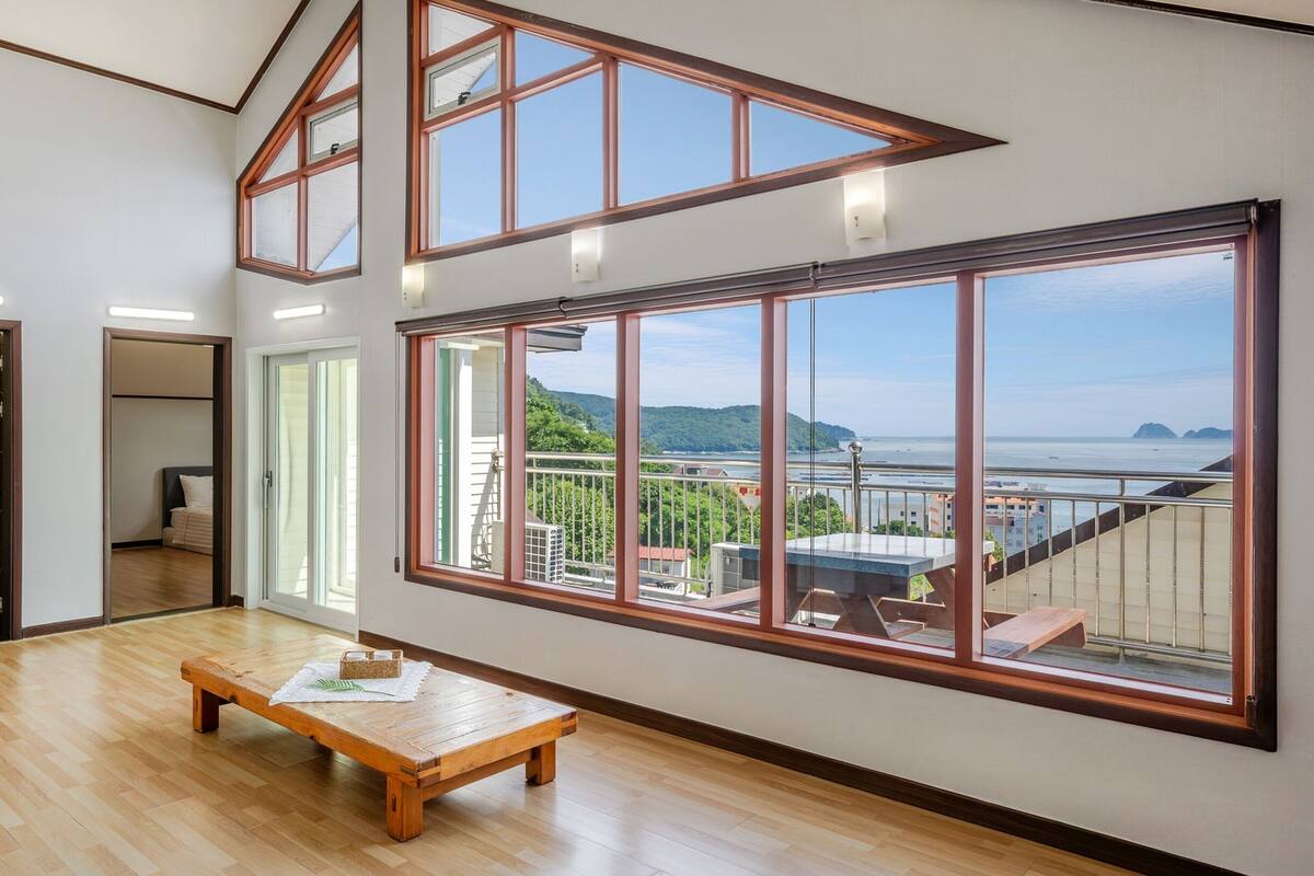 A spacious room with large windows offering a view of the sea and hills. A wooden bench and a small table are in the center.