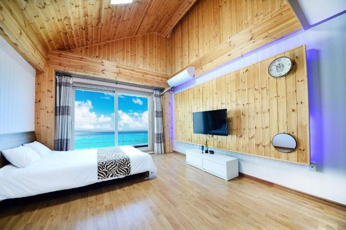 Une chambre avec un plafond en bois, un lit, une télévision, une fenêtre donnant sur l’océan et une horloge fixée au mur.