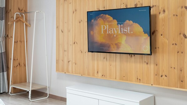 A room with a wooden wall, a white TV mounted on it displaying a playlist, a white cabinet, and a metal hanger stand. - Donghae Red Roof Pension (Donghae)