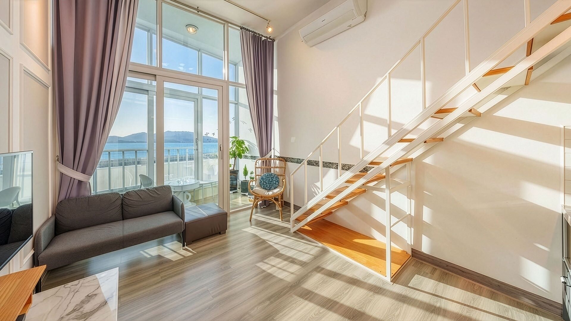 A modern living room with a sofa, a dining table, a staircase, and large windows with curtains.