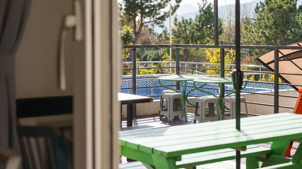 Un balcón con vista a una piscina de azulejos azules, mobiliario exterior verde y árboles al fondo.